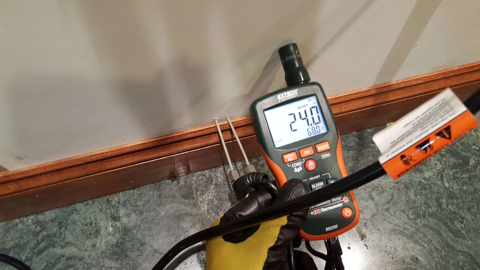 Extech pin-type moisture meter showing 24 percent reading at baseboard indicating wet wall
